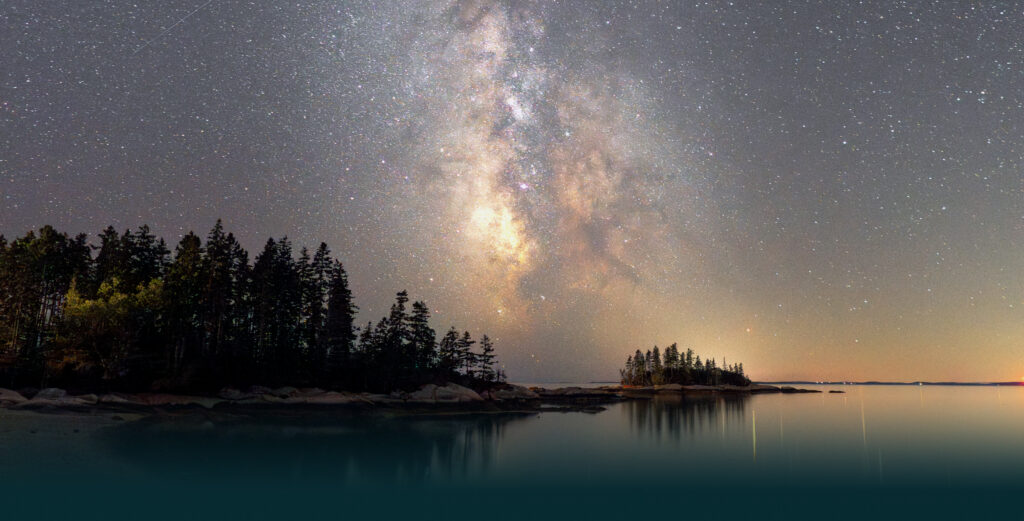 Milky Way stretching across a star-filled night sky over a calm lake with a dark treeline silhouette at the shore.