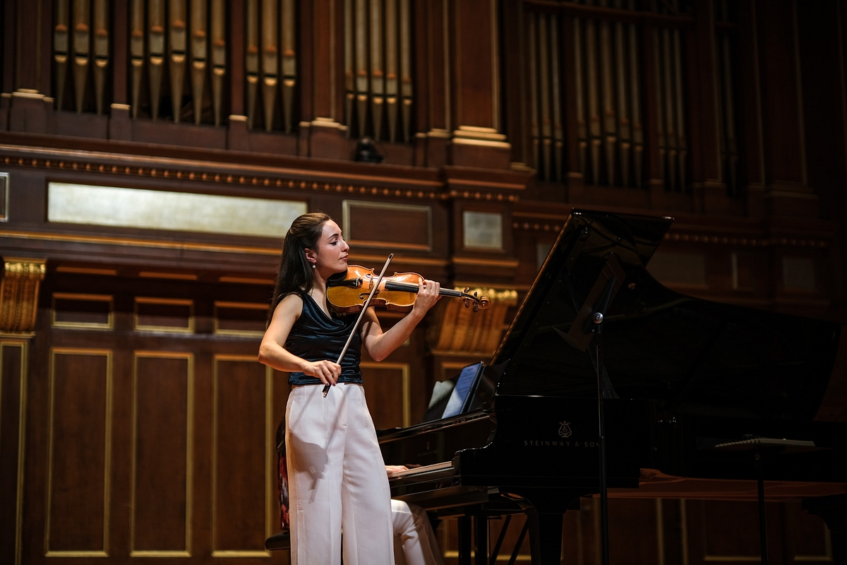 Female violinist performing on a stage with a grand piano in a wood-paneled concert hall.