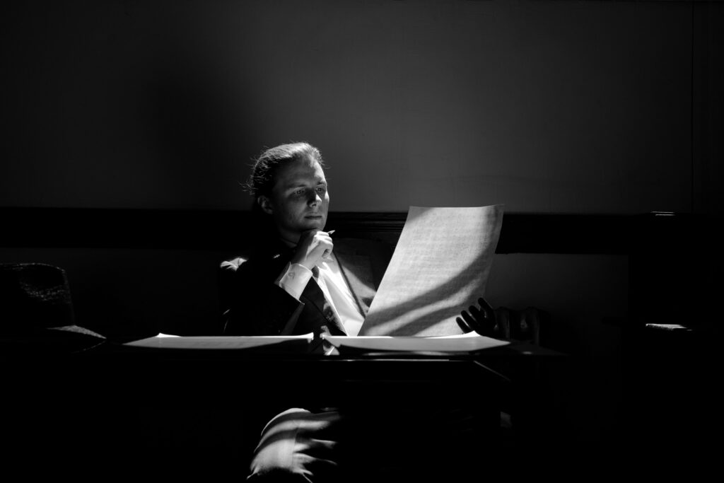 Black-and-white photo of a man in a suit seated at a desk, studying a large sheet of paper with a thoughtful pose under dramatic lighting.